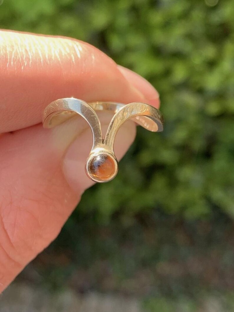 May include: A gold-toned ring featuring a V-shaped band that splits into two arms, meeting at a central point where a small, round, amber-colored gemstone is set. The metal has a smooth, polished finish, and the design creates a distinct arching shape.