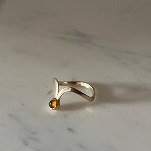 May include: A gold-toned ring featuring a curved, open-ended band design that tapers to a point. One end of the band is accented with a single, small, round, amber-colored gemstone set in a smooth, polished metal bezel setting.