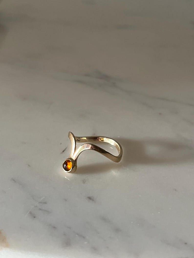 May include: A gold-toned ring featuring a curved, open-ended band design that tapers to a point. One end of the band is accented with a single, small, round, amber-colored gemstone set in a smooth, polished metal bezel setting.