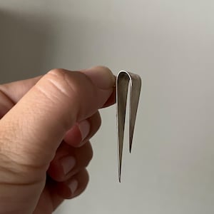 May include: A silver-toned metal earring featuring a long, slender, folded design. The piece consists of two pointed, tapering prongs that meet at a rounded top, creating a sharp, elongated silhouette with a smooth, matte metallic finish.
