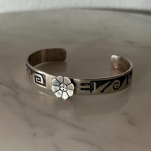 May include: Silver cuff bracelet with a floral design and geometric patterns. The bracelet features a flower in the center and decorative lines and shapes along the band. The open cuff design allows for easy wear.