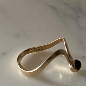 May include: A gold ring with a wavy, open-ended band design featuring a single dark red stone set at one terminal. The interior of the band displays a series of small, engraved hallmarks indicating the metal purity and origin.