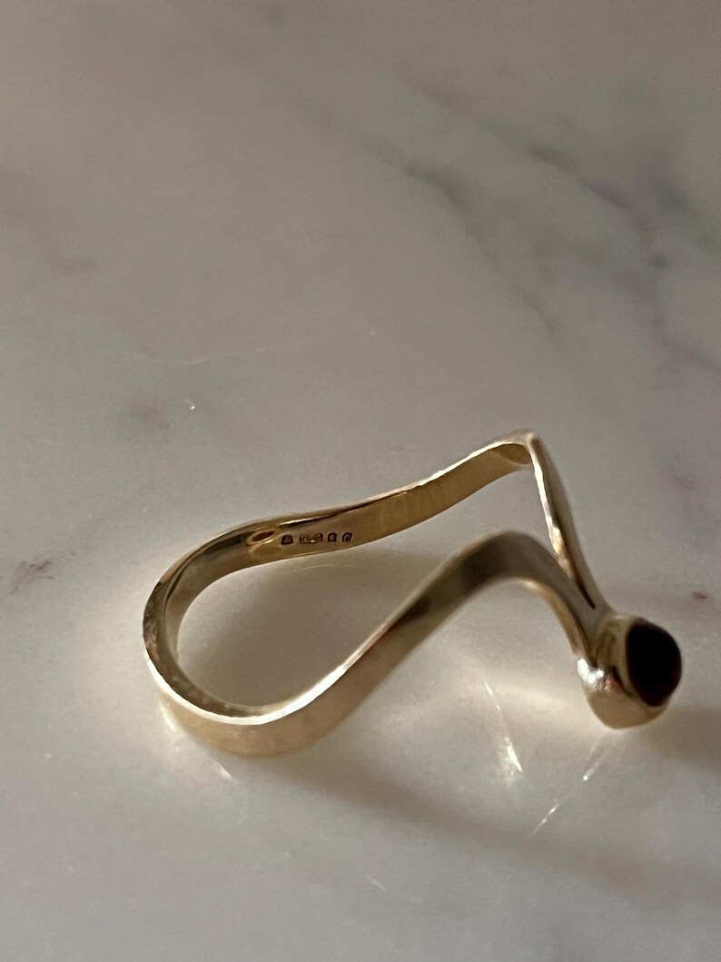 May include: A gold ring with a wavy, open-ended band design featuring a single dark red stone set at one terminal. The interior of the band displays a series of small, engraved hallmarks indicating the metal purity and origin.
