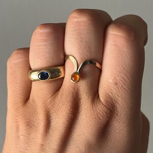 May include: Two gold rings are displayed. One ring has a blue oval stone, and the other has a curved design with an orange stone. The rings are on a hand, showcasing the jewelry's design and color.