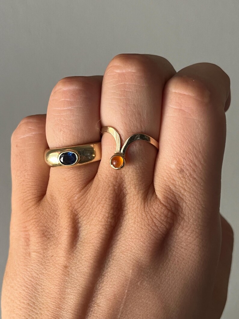 May include: Two gold rings are displayed. One ring has a blue oval stone, and the other has a curved design with an orange stone. The rings are on a hand, showcasing the jewelry's design and color.