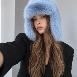 May include: A light blue faux fur trapper hat with a thick, fluffy texture and ear flaps that extend down the sides. The hat features a dense, soft pile that covers the crown and frames the face with a voluminous, plush appearance.
