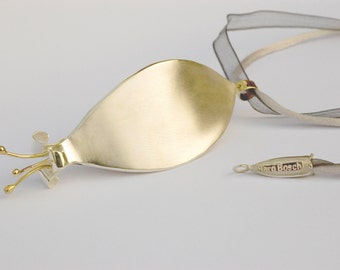 Puede incluir: Un collar de plata y oro con un gran colgante ovalado liso y un delicado detalle en forma de flor. El collar está unido a una cinta fina gris con una pequeña etiqueta plateada que dice "Haro Bosch".