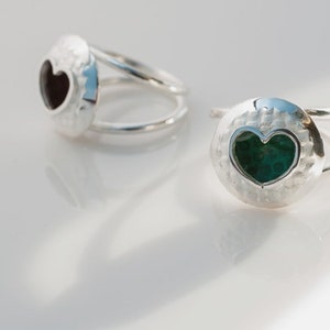 May include: Two silver rings with a heart-shaped cutout. One ring has a black heart-shaped stone, and the other has a green heart-shaped stone.