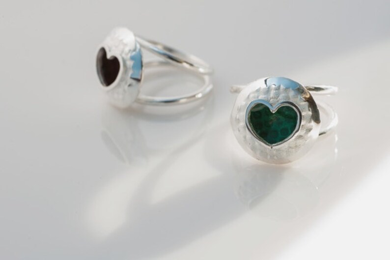 May include: Two silver rings with a heart-shaped cutout. One ring has a black heart-shaped stone, and the other has a green heart-shaped stone.