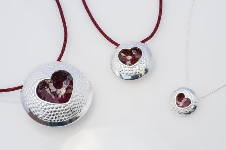 May include: Three silver necklaces with red heart pendants. The pendants are round and have a textured surface. The necklaces are of different lengths.