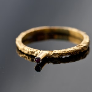 Op de afbeelding: Een gouden ring met een getextureerde band en een kleine rode edelsteen gezet in een gouden klauw.