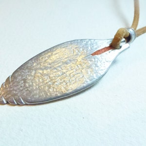Puede incluir: Un colgante de plata con forma de hoja y superficie texturizada. El colgante está unido a un cordón de cuero marrón.