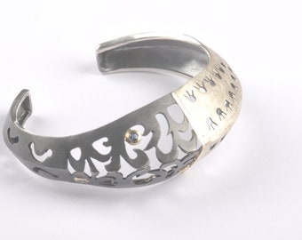 May include: A silver and gold bracelet with a cutout design and blue gemstones. The bracelet is open and has a textured surface.