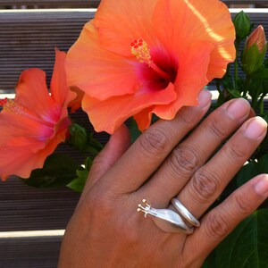 May include: A silver ring with a gold snail design. The ring is worn on a hand with orange hibiscus flowers in the background.