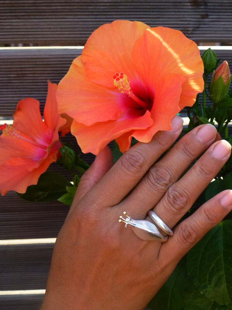 May include: A silver ring with a gold snail design. The ring is worn on a hand with orange hibiscus flowers in the background.