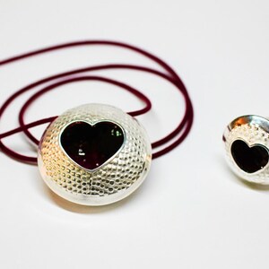 May include: A silver necklace and ring set with a heart-shaped cutout and a black background. The necklace has a burgundy cord.
