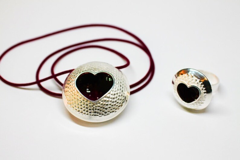 May include: A silver necklace and ring set with a heart-shaped cutout and a black background. The necklace has a burgundy cord.