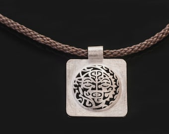 May include: A silver pendant necklace with a square pendant featuring a detailed cutout design of a face. The pendant is attached to a brown cord.
