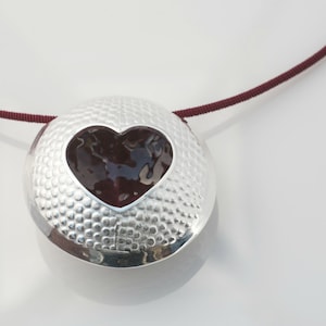 May include: A silver pendant necklace with a textured surface and a heart-shaped cutout filled with a dark red material. The necklace is on a maroon cord.