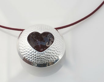 May include: A silver pendant necklace with a textured surface and a heart-shaped cutout filled with a dark red material. The necklace is on a maroon cord.
