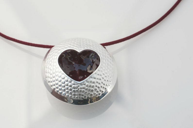 May include: A silver pendant necklace with a textured surface and a heart-shaped cutout filled with a dark red material. The necklace is on a maroon cord.