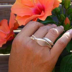 May include: A silver ring with a gold accent in the shape of a snail. The ring is worn on a person's finger, with a pink hibiscus flower in the background.