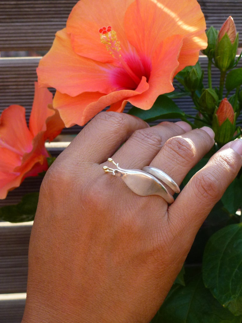 May include: A silver ring with a gold accent in the shape of a snail. The ring is worn on a person's finger, with a pink hibiscus flower in the background.