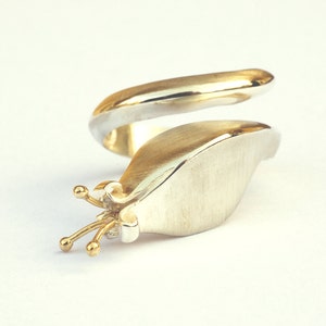 May include: A silver and gold ring with a unique design. The ring is shaped like a snail with a gold shell and a silver body. The snail has a small gold crown on its head.