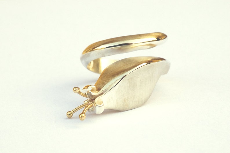 May include: A silver and gold ring with a unique design. The ring is shaped like a snail with a gold shell and a silver body. The snail has a small gold crown on its head.