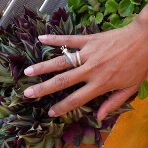 May include: A silver ring with a crown design on the top. The ring is on a person's finger and the background is a green plant.