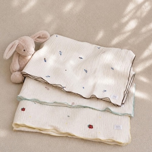 May include: A beige plush rabbit toy sits next to two folded off-white muslin blankets. The top blanket features small blue floral embroidery and a dark brown edge. The bottom blanket displays tiny strawberry and yellow flower motifs with a yellow edge.
