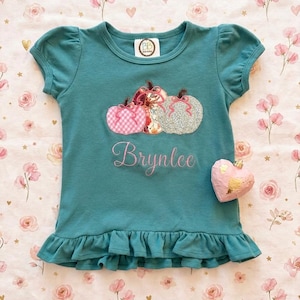 Puede incluir: Una camiseta de manga corta color verde azulado con dobladillo de volantes, que presenta un diseño bordado de tres calabazas en patrones de cuadros rosas, flores y plata. El nombre Brynlee está bordado en letra cursiva rosa.
