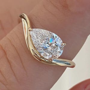 May include: A gold ring featuring a single pear-shaped clear gemstone set in a bypass-style band. The band curves around the base of the stone, creating a fluid, asymmetrical design that highlights the faceted cut of the central stone.