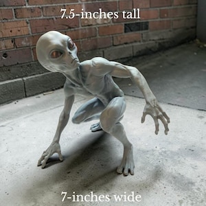 May include: A light gray alien figurine in a crouching pose with large dark eyes and elongated limbs. The figure measures 7.5-inches tall and 7-inches wide, as indicated by the text overlay. The alien has a smooth, detailed skin texture.