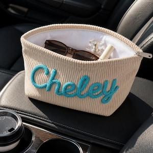 May include: A beige ribbed fabric pouch with a zipper closure, featuring the name Cheley embroidered in bright blue yarn on the front. The open pouch contains a pair of dark-rimmed sunglasses, a small glass perfume bottle, and a lipstick tube.