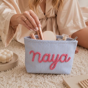 May include: A light blue and white pinstriped fabric pouch featuring the name naya embroidered in raised pink yarn on the front. The open top reveals various cosmetic items including a rose gold tube and a small glass perfume bottle.