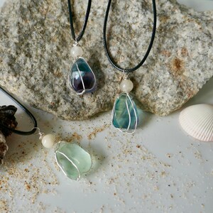 Può includere: Tre collane con pietre di fluorite grezze e irregolari nelle tonalità del viola, blu e verde chiaro. Ogni pietra è fissata in un avvolgimento di filo color argento con una piccola perla rotonda color bianco sporco su un cordino nero.