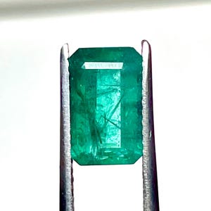 Puede incluir: Una esmeralda rectangular de tono verde vibrante con inclusiones internas visibles, sostenida firmemente por unas pinzas de metal plateado. La piedra preciosa presenta un corte facetado que refleja la luz sobre su superficie translúcida.