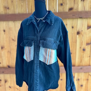 May include: A black denim button-down shirt featuring a classic collar, long sleeves with buttoned cuffs, and two chest pockets. The lower portion of each chest pocket is accented with a vertical striped pattern in shades of blue, orange, and white.