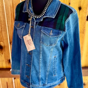May include: A blue denim jacket featuring dark green and navy blue plaid panels on the upper chest and shoulders. The jacket has a collared neckline, metal button closures, two chest pockets, and a beige paper tag with the text The Reclaimed Cowgirl.