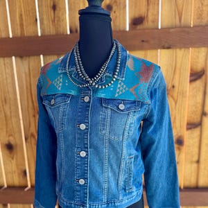 May include: A blue denim jacket featuring a turquoise yoke with a brown and tan geometric pattern across the shoulders and chest. The jacket has a classic collar, metal button closures, two chest pockets with flaps, and long sleeves.