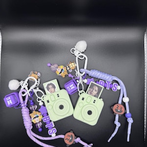 May include: Two light green instant camera keychains with purple braided lanyards. Each features a small photo of a person, a white pom-pom, a purple cube charm with the text BTS, and various character charms including a brown bear and a red creature.