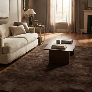 May include: A large rectangular area rug in a deep dark brown color with a subtle textured finish, positioned on a wooden floor in a living room setting that includes a light beige sofa, a low wooden coffee table, and sheer window curtains.