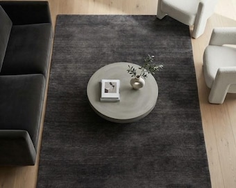 May include: A large rectangular area rug in a dark charcoal gray tone with a subtle horizontal texture. The rug is positioned on a light wood floor, serving as a base for a round light gray coffee table, a small white vase, and a book.