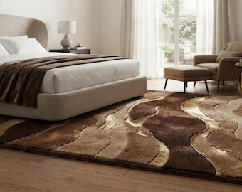 May include: A rectangular area rug featuring a multi-tonal abstract pattern of wavy, flowing lines in shades of dark brown, medium brown, tan, and cream. The rug has a textured, plush pile surface and is positioned on a light-colored wooden floor.
