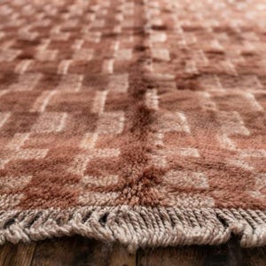 Könnte beinhalten: Ein rechteckiger Teppich mit einem geometrischen Muster in tiefem Terrakotta und Creme mit einem sich wiederholenden Blockdesign. Der Teppich hat eine dicke, hochflorige Textur und ist an der Unterkante mit einer Franse aus cremefarbenem Garn versehen.