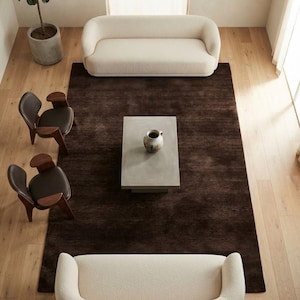 May include: A living room arrangement featuring two cream-colored curved sofas, two dark brown wooden chairs with black seats, a rectangular gray coffee table with a ceramic vase, and a large dark brown area rug on light wood flooring.