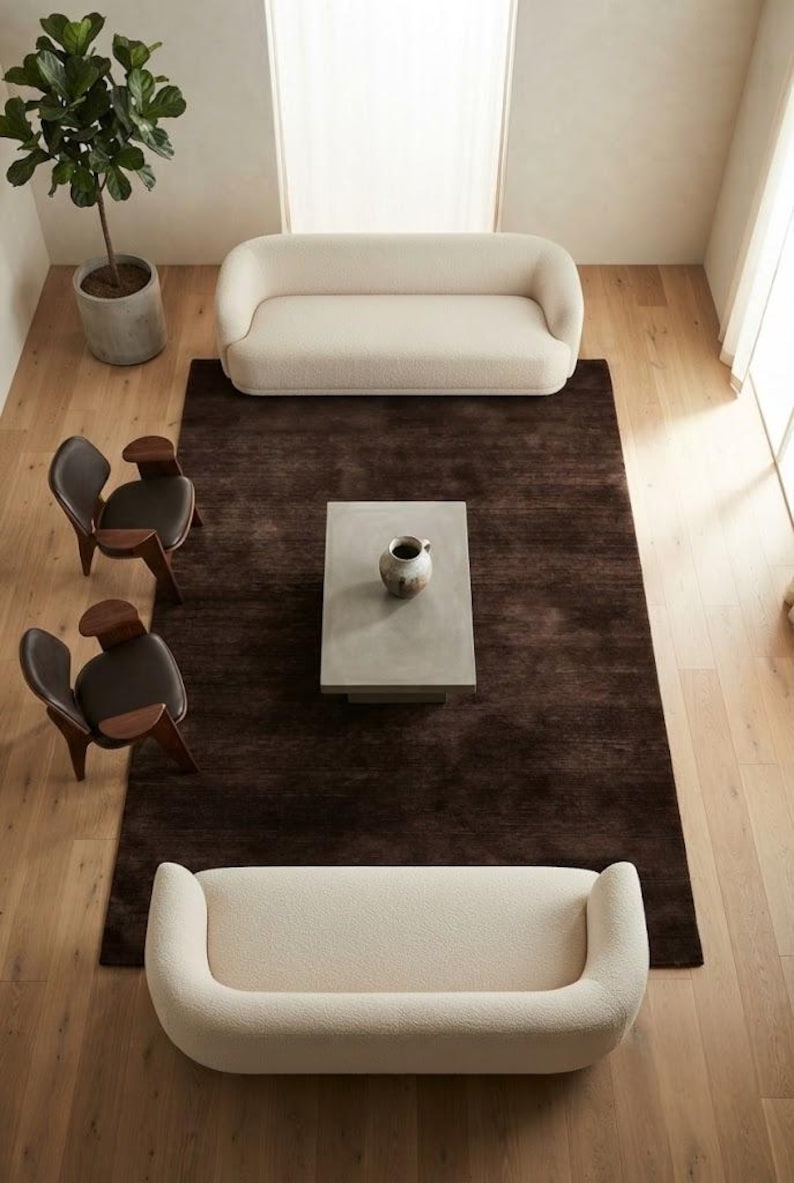 May include: A living room arrangement featuring two cream-colored curved sofas, two dark brown wooden chairs with black seats, a rectangular gray coffee table with a ceramic vase, and a large dark brown area rug on light wood flooring.