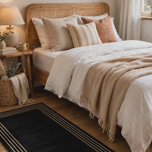 Könnte beinhalten: Ein rechteckiger schwarzer Teppich mit einer dreifachen Umrandung in Hellbraun oder Gold. Der Teppich liegt auf einem Holzboden vor einem Bett mit einer hellen Leinenbettdecke, einer strukturierten beigen Tagesdecke und verschiedenen Kissen.