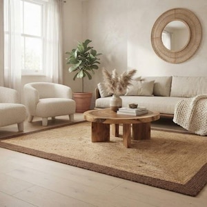 Könnte beinhalten: Ein Wohnzimmer mit einem rechteckigen Juteteppich mit dunkelbraunem Rand, zwei cremefarbenen, strukturierten Sesseln, einem hellen Sofa mit einer grob gestrickten Decke, einem runden Couchtisch aus Holz und einem runden, geflochtenen Wandspiegel.